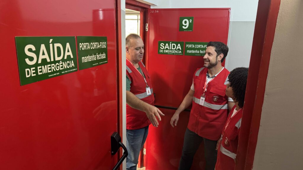 Brigadistas junto a uma porta corta-fogo vermelha com sinalização de "Saída de Emergência", a demonstrar procedimentos nas rotas de evacuação do prédio.