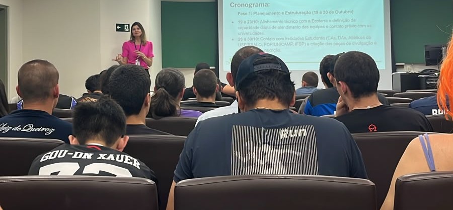 Uma mulher vestida de cor-de-rosa faz uma apresentação junto a um ecrã de projeção, enquanto um auditório com dezenas de estudantes assiste a partir dos lugares sentados.