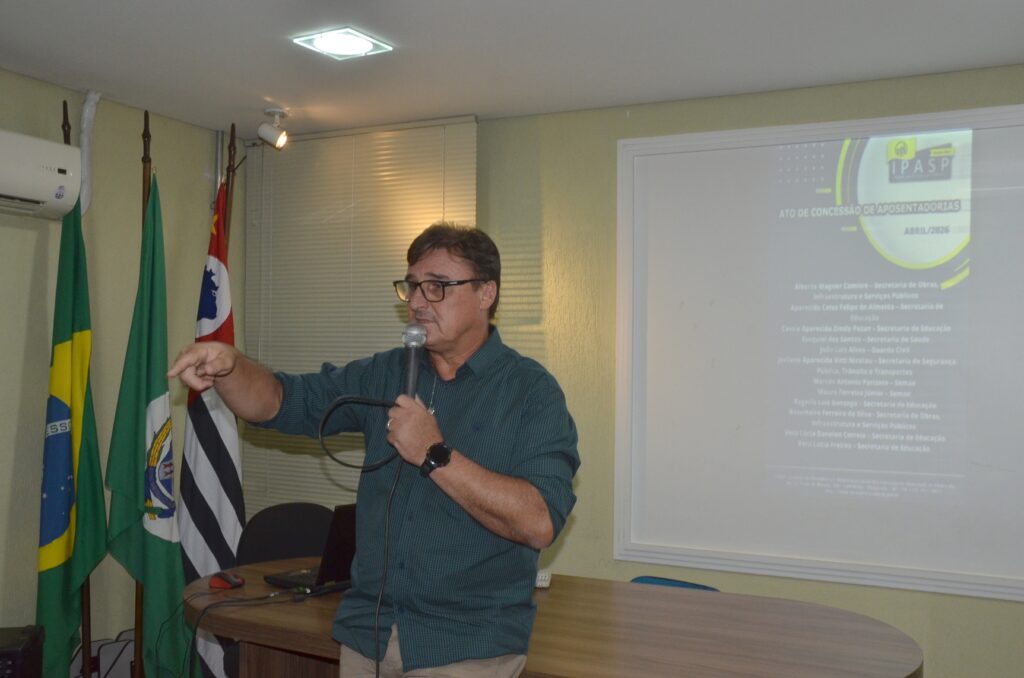 Carlinhos Schiavon, presidente do Ipasp, fala ao microfone e gesticula durante uma apresentação em auditório, tendo ao lado as bandeiras do Brasil, do Estado de São Paulo e do município, e ao fundo uma projeção do instituto