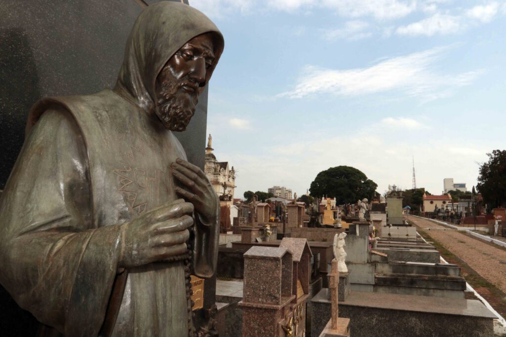 Apresenta um plano aproximado de uma estátua de bronze de uma figura religiosa (São Francisco de Assis) num cemitério, com uma perspetiva alargada ao fundo que mostra as alamedas e diversos jazigos sob um céu parcialmente nublado.