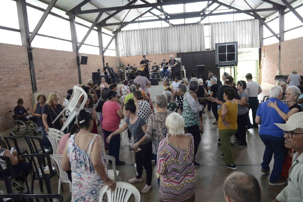 Salão amplo com estrutura metálica e paredes de tijolo à vista repleto de idosos a dançar, com uma banda a tocar ao vivo num palco ao fundo.