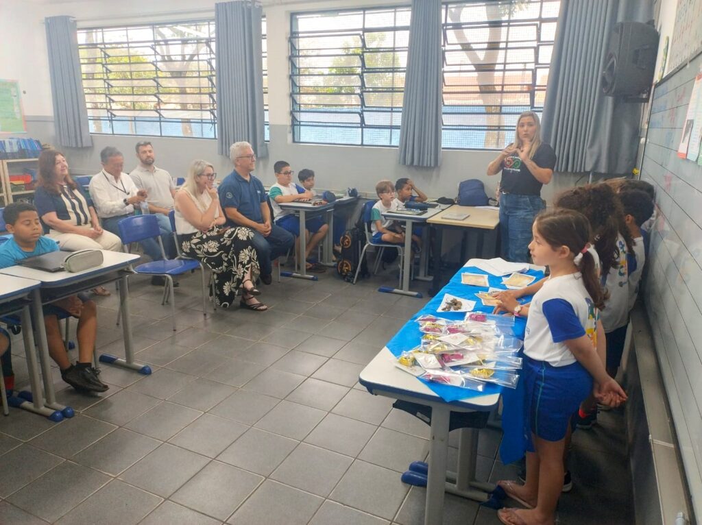 Alunos em pé junto a uma mesa com materiais expostos apresentam um trabalho na sala de aula, enquanto uma mulher em pé traduz ou comunica em Língua Gestual e um grupo de adultos e estudantes assiste.