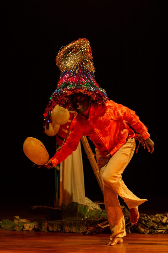 Um artista performativo em trajes coloridos e um grande chapéu cónico decorado com fitas reluzentes dança no palco, segurando um instrumento artesanal sob um fundo escuro.