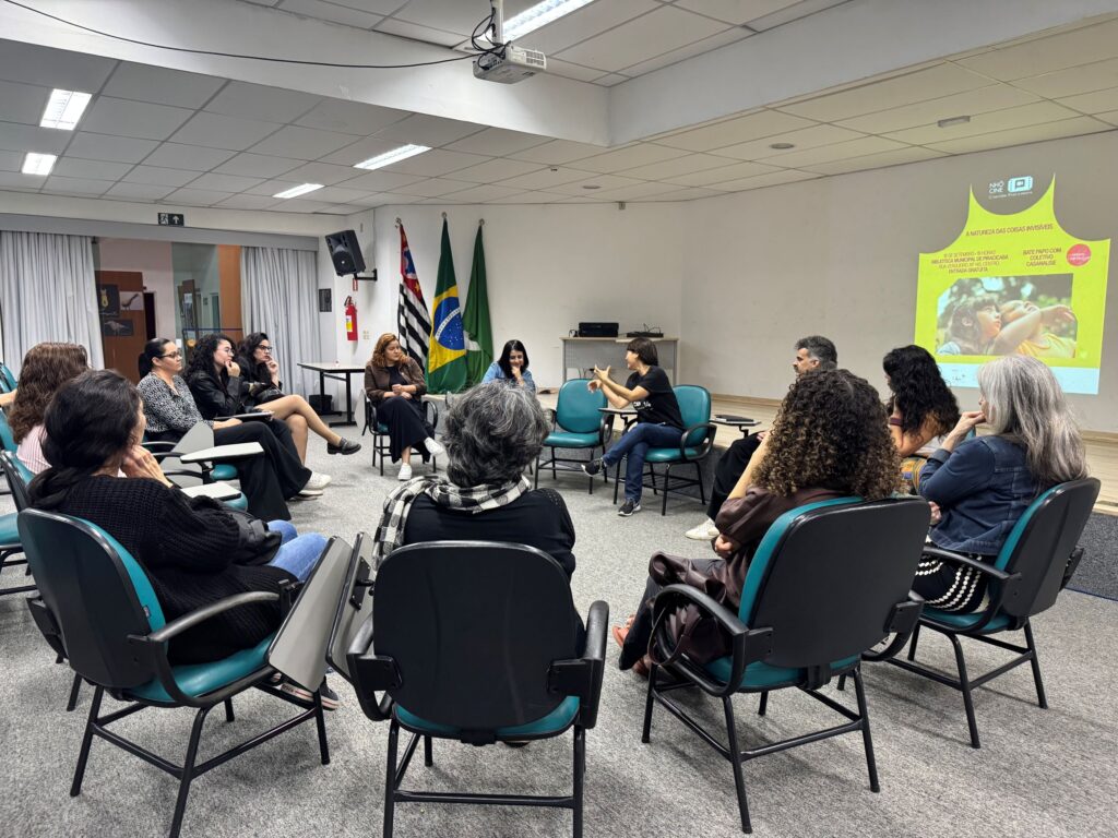 Grupo de pessoas dispostas em círculo numa sala de reuniões durante um debate ou roda de conversa do Nhô Cine, com um projetor a exibir uma apresentação na parede.