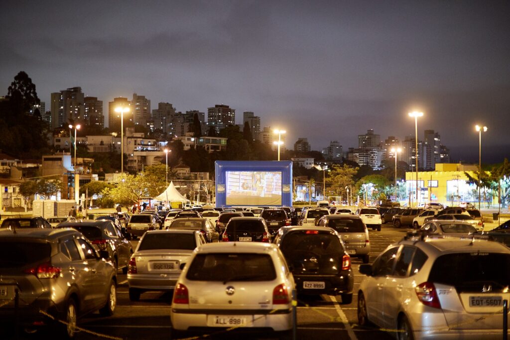 Vista panorâmica e noturna de um parque de estacionamento repleto de automóveis virados para um grande ecrã insuflável de cinema drive-in (Cine Autorama). Ao fundo, avista-se a paisagem urbana iluminada com edifícios e postes de iluminação.