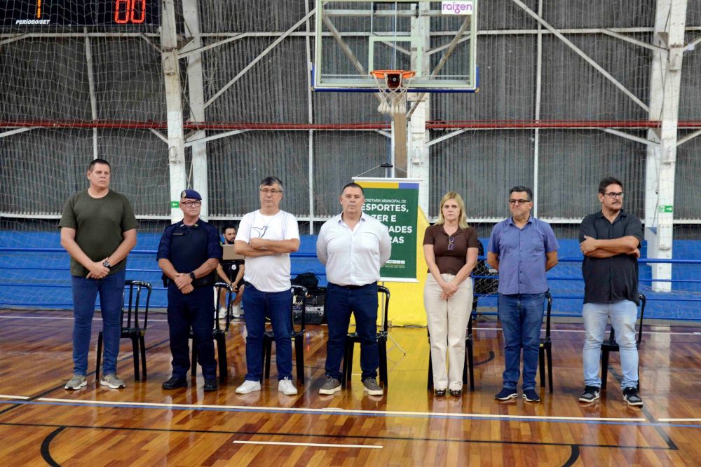 Autoridades e organizadores do evento posam lado a lado na quadra em frente à tabela de basquetebol durante a cerimónia de abertura.