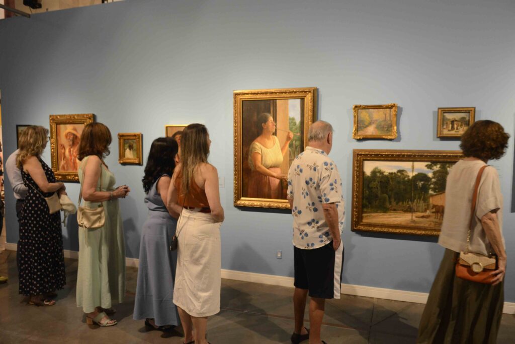 Visitantes numa galeria de arte a observar várias pinturas com temas caipiras expostas ao longo de uma parede azul.