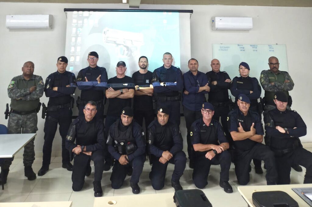 Grupo de guardas civis municipais e instrutores fardados posam para uma foto oficial numa sala de formação, dispostos em duas fileiras (uma de pé e outra ajoelhada), tendo ao fundo um ecrã de projeção a exibir a imagem de uma arma de fogo.