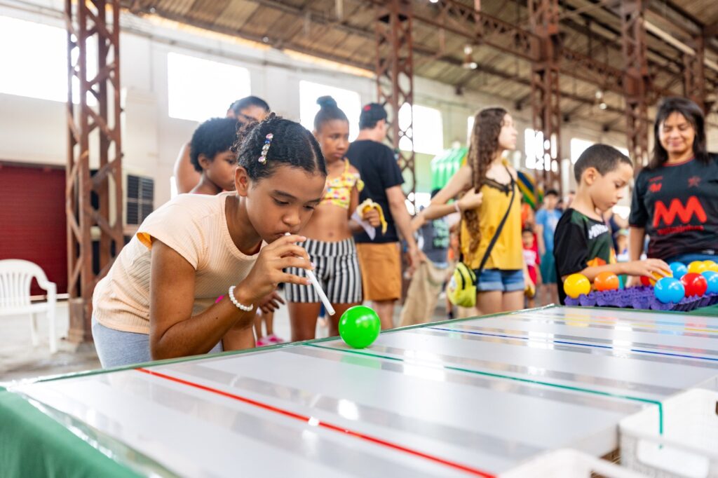 Menina a soprar uma bola verde através de uma palhinha num jogo de mesa recreativo, rodeada por outras crianças no Engenho Central.
