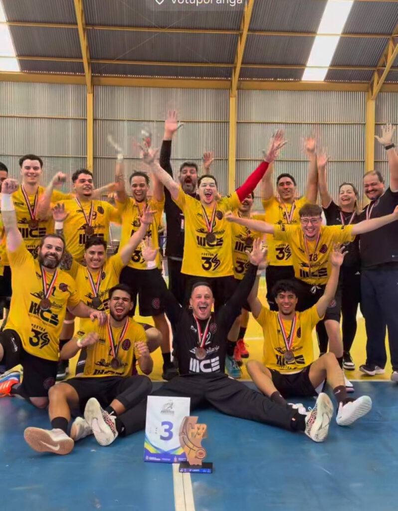 Uma equipe masculina de handebol posa feliz para a câmera no chão de um ginásio coberto. Eles vestem uniformes amarelos e pretos (da SELAM Piracicaba), exibem medalhas e comemoram a terceira colocação, com um troféu e uma placa com o número '3' à frente