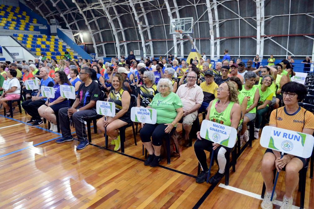 Dezenas de participantes de diferentes delegações estão sentados em cadeiras dispostas no pavilhão, segurando placas com os nomes dos seus respetivos grupos.