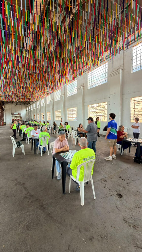 Visão ampla do espaço interior com teto decorado por fitas coloridas penduradas. Vários idosos estão dispostos em mesas individuais a jogar damas durante a competição.