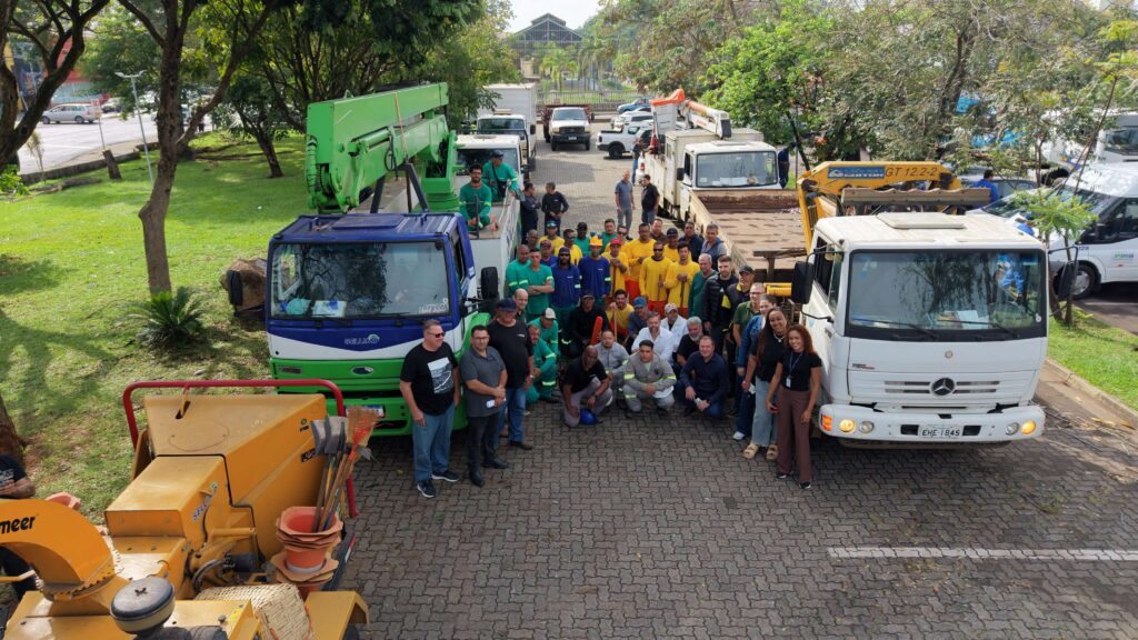 Foto de grupo com as equipas de trabalho e gestores reunidos num parque de estacionamento, ladeados por camiões de manutenção e equipamento de trituração de ramos.