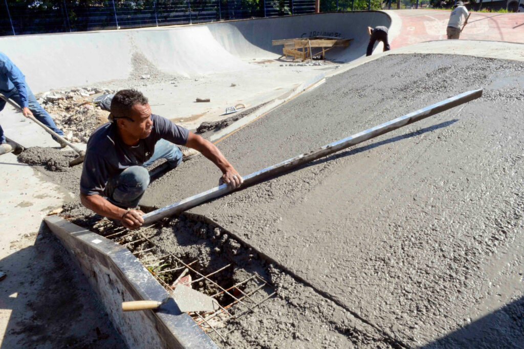 Em plano aproximado, um pedreiro ajoelhado utiliza uma régua de alumínio para alisar e nivelar o betão fresco numa das rampas de uma pista de skate em obras. Ao fundo, outros trabalhadores auxiliam nas obras da pista.