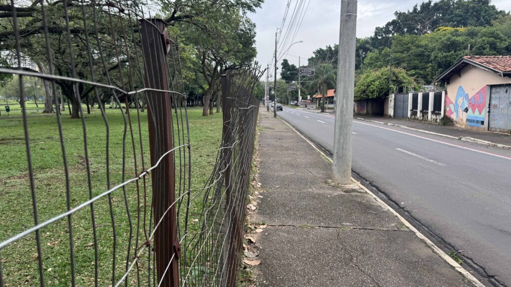 Vista em perspetiva de uma rede metálica danificada e torta ao longo de um passeio pedonal, dividindo a via urbana de uma área verde com árvores.