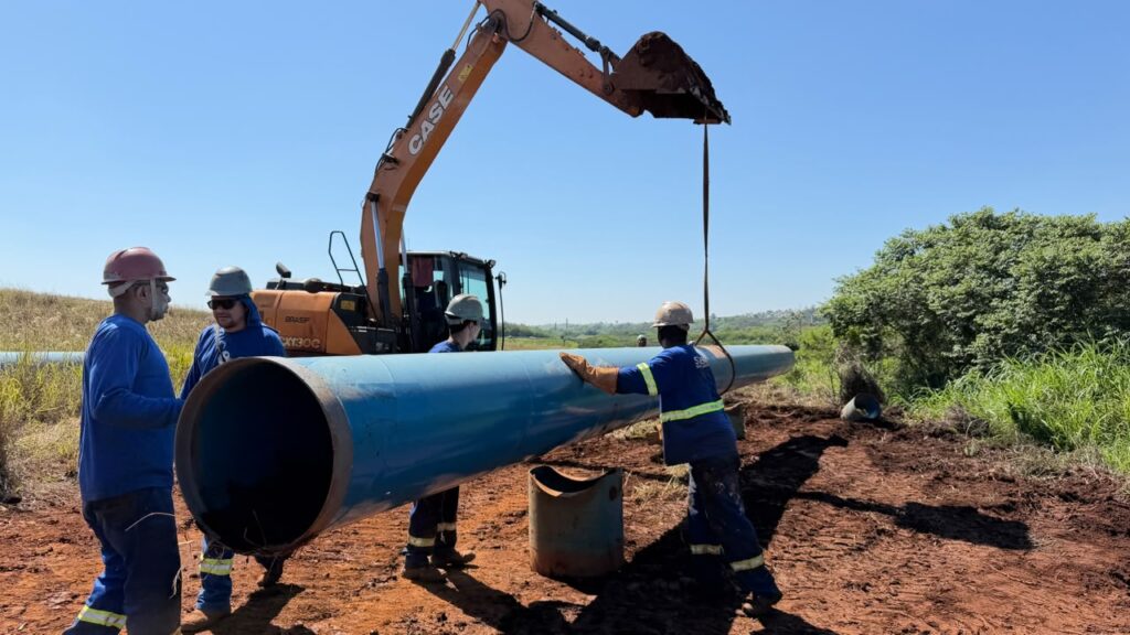 Trabalhadores utilizam uma escavadeira para içar e posicionar um tubo de grande porte durante a construção de uma adutora. A atividade ocorre em terreno aberto sob céu limpo.