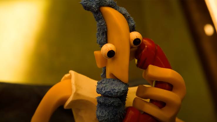 Cena da animação em stop-motion Contra Filé, destacando uma personagem amarela feita de massa de modelar com olhos arregalados e detalhes azuis na cabeça e barba, a atender um telefone vermelho.