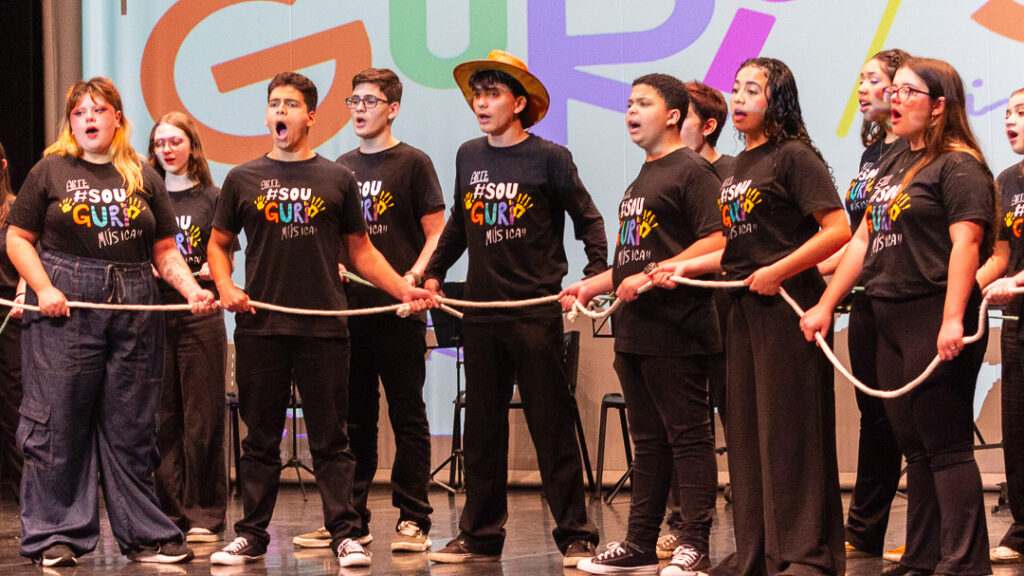 O grupo continua a apresentação musical, em pé e cantando com empolgação, enquanto todos seguram uma corda branca que se estende por todo o grupo. Eles vestem camisetas pretas com a frase "#Sou Guri Música"