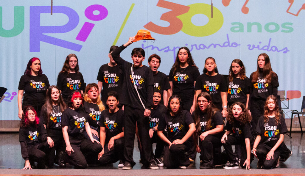 Um grupo de crianças e adolescentes do Projeto Guri se apresenta em um palco, cantando e fazendo poses, com um deles levantando um chapéu. Ao fundo, um telão comemora os "30 anos inspirando vidas"