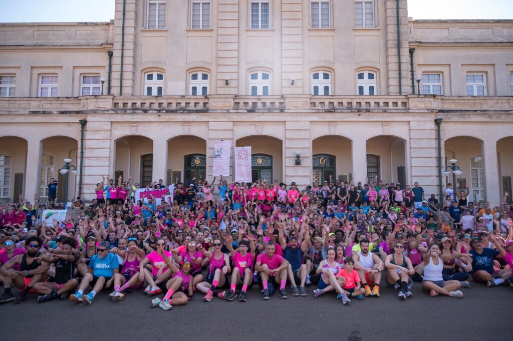Um grande grupo de corredores e participantes da Corrida Corta Mato do Outubro Rosa, muitos deles vestidos com camisolas cor-de-rosa, posa reuniu-se sentado e de pé em frente à fachada histórica de um edifício imponente para uma foto coletiva.