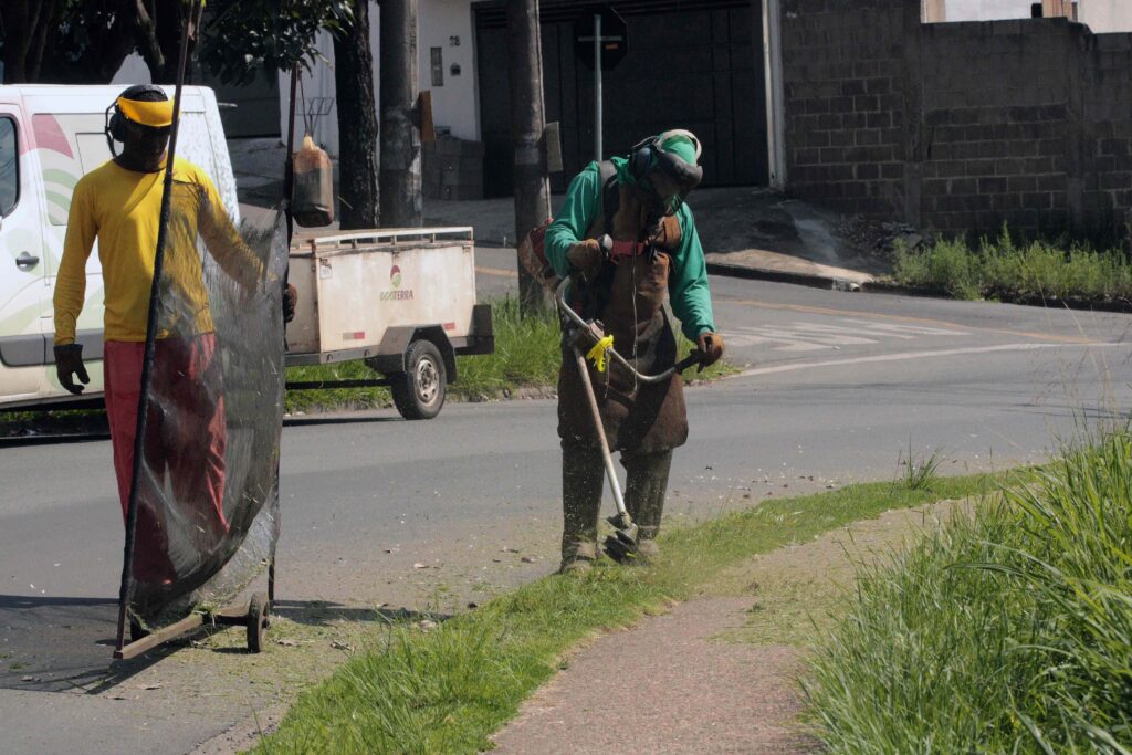 Um operador utiliza uma roçadeira motorizada no canteiro/calçada, enquanto outro trabalhador segura uma proteção móvel para evitar que pedras ou detritos atinjam os veículos.