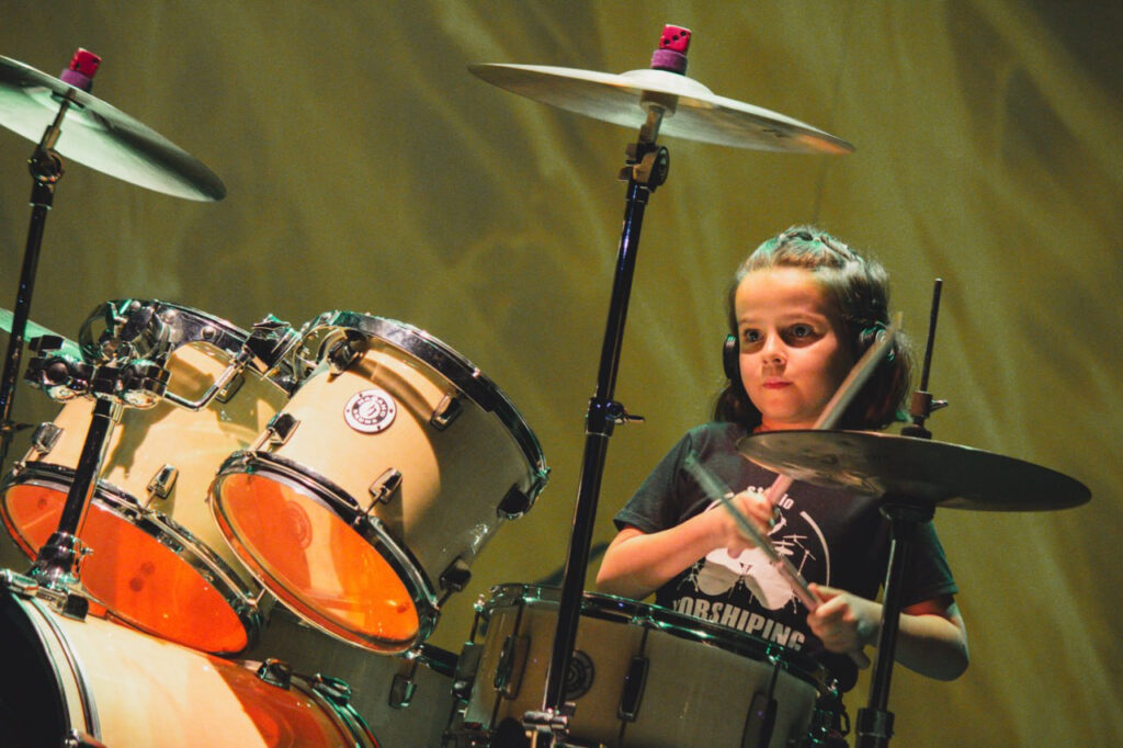 Uma criança pequena com auscultadores de proteção segura nas baquetas e toca numa bateria acústica durante uma apresentação em palco.