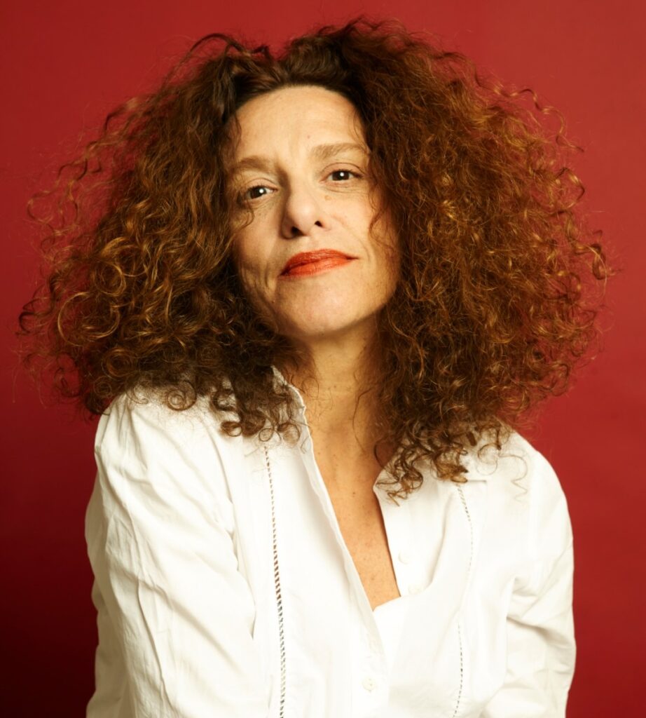 Retrato da atriz Yara de Novaes, posando para a câmera vestindo uma camisa branca contra um fundo vermelho vivo