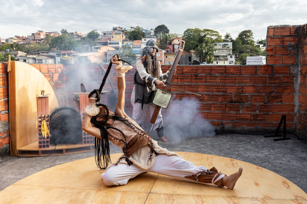 Duas artistas apresentam o espetáculo híbrido "Mary para Mary" em uma laje com uma favela ao fundo. Uma delas faz uma pose acrobática no chão enquanto a outra toca um instrumento de cordas inventado