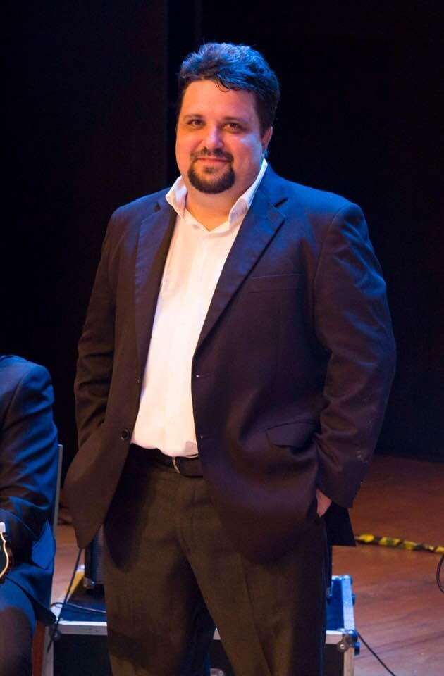Um homem de fato escuro e camisa branca posa sorridente de pé num palco com iluminação cénica ao fundo.