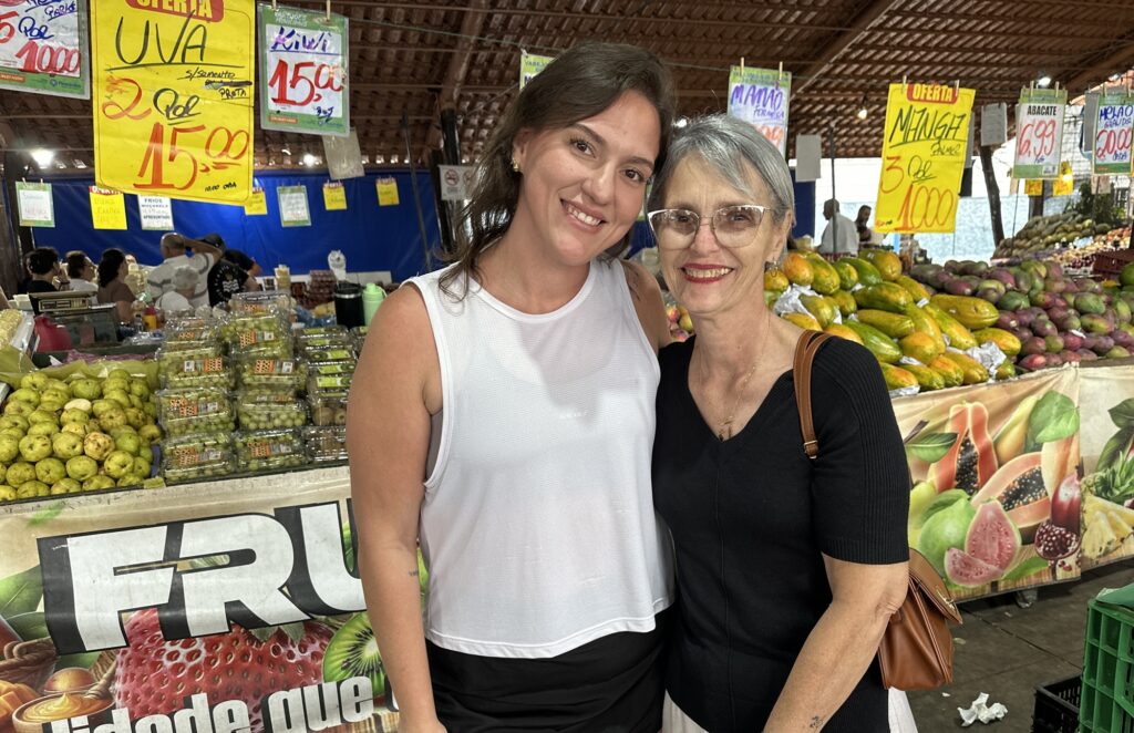 Thais e Viviane, filha e mãe, sorriem abraçadas em frente às bancas de frutas no Varejão da Paulista.