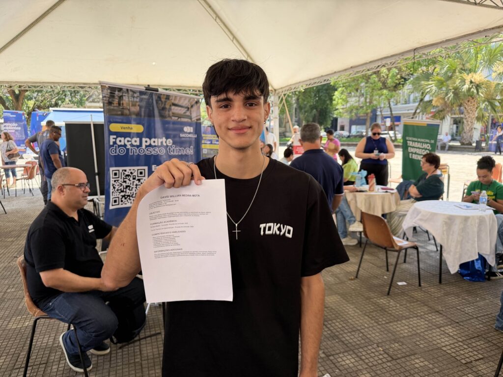 O jovem David William Medina posa para a foto segurando o seu currículo em papel diante das tendas de atendimento.