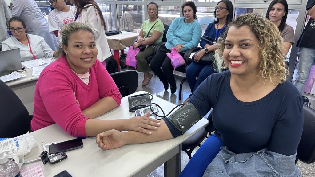 Mostra Deise Arruda, motorista de 36 anos, a aferir a pressão arterial e a realizar exames de saúde numa mesa de atendimento com outras pessoas ao fundo.