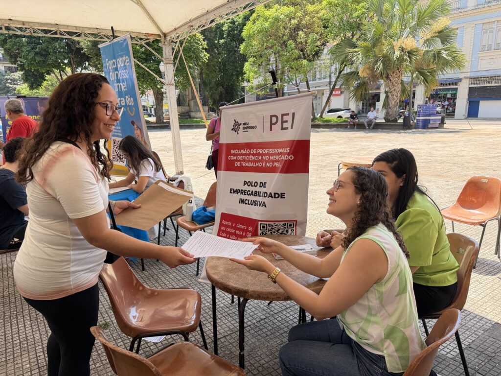Janaina Schiavinato em pé entrega uma folha impresso a uma atendente sentada numa banca do Polo de Empregabilidade Inclusiva (PEI).