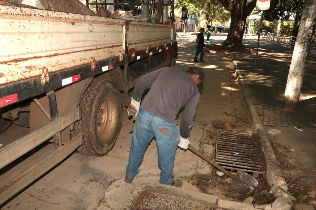 Mostra um trabalhador debruçado junto à traseira de um caminhão de caixa aberta, a utilizar uma ferramenta manual para desobstruir e limpar uma boca de lobo (bueiro) no meio-fio de uma rua pavimentada. Ao fundo, avista-se outro trabalhador numa zona arborizada.