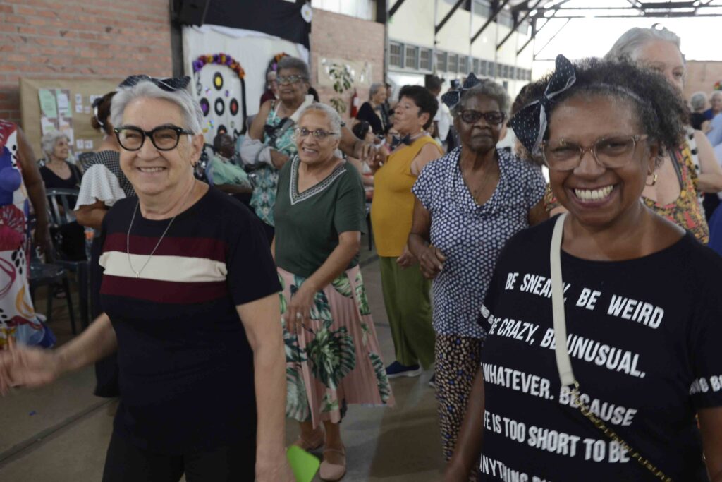 Várias mulheres idosas a sorrir enquanto dançam em grupo no salão, vestidas com roupas casuais e lacos de fita na cabeça.