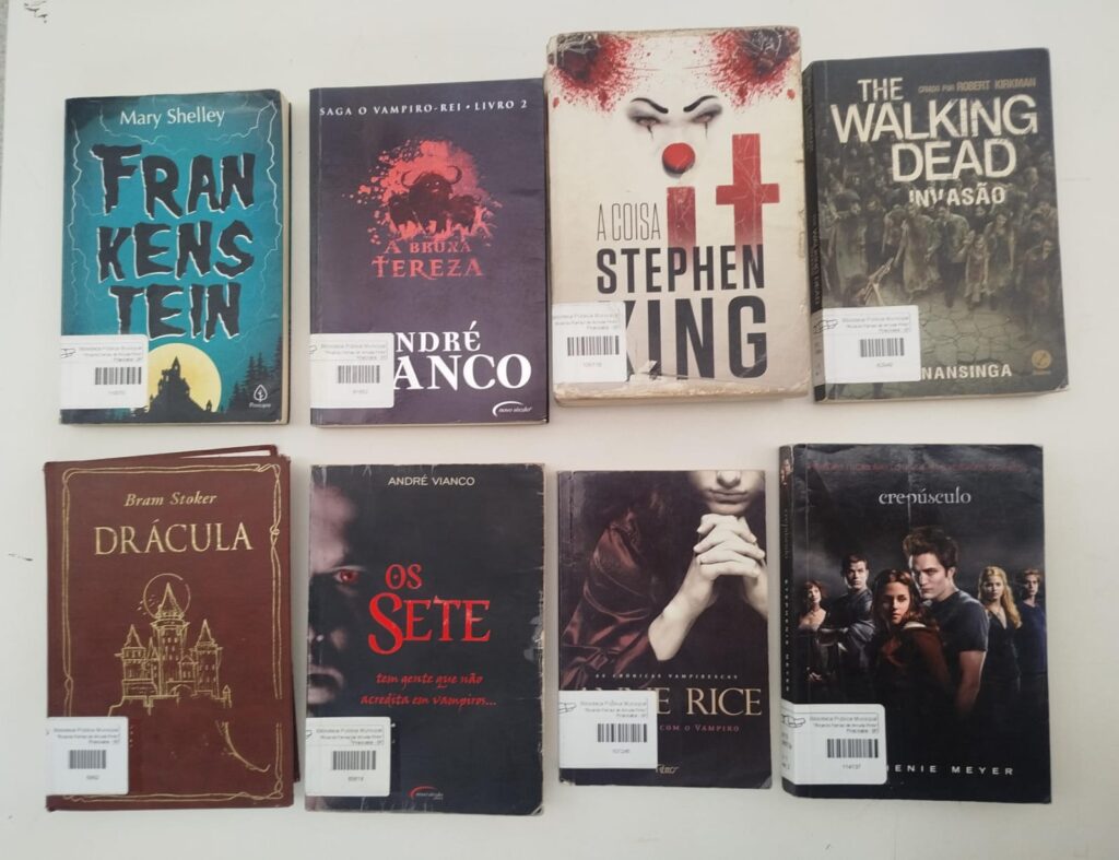 Oito livros com temática de suspense, terror e vampiros do acervo da biblioteca dispostos sobre uma superfície clara, incluindo títulos como Frankenstein, Drácula, IT: A Coisa, Os Sete e Crepúsculo.