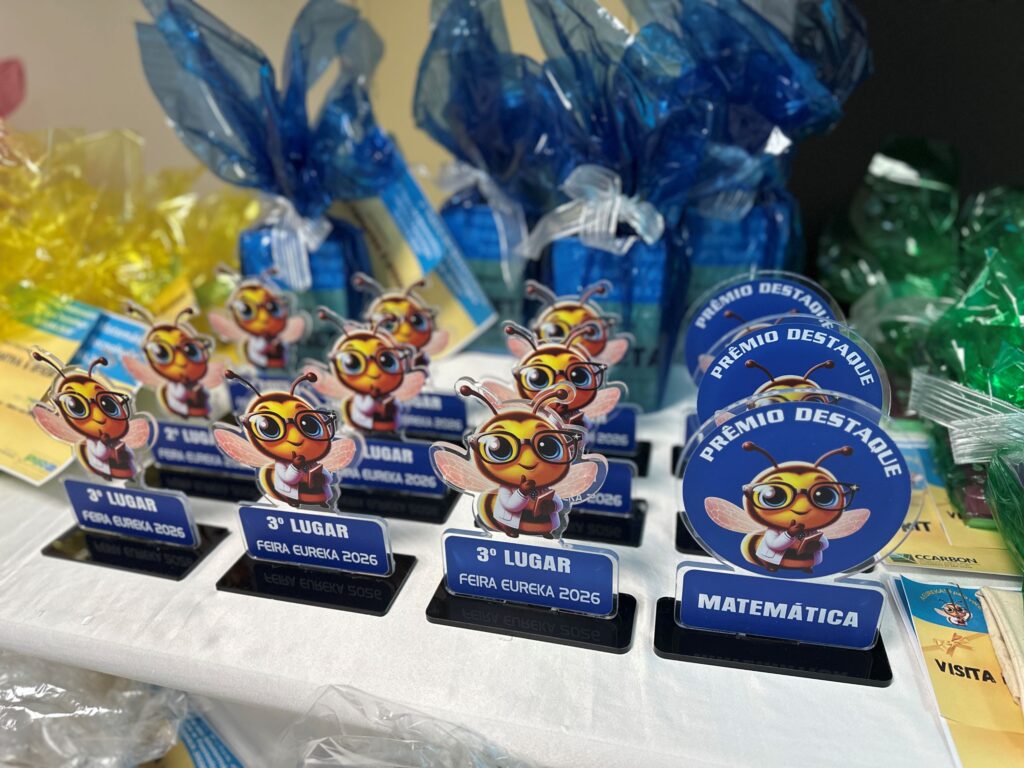 Vários troféus e prémios alinhados numa mesa, decorados com uma mascote em forma de abelha e etiquetas indicando categorias e posições como o 3.º lugar e matemática.