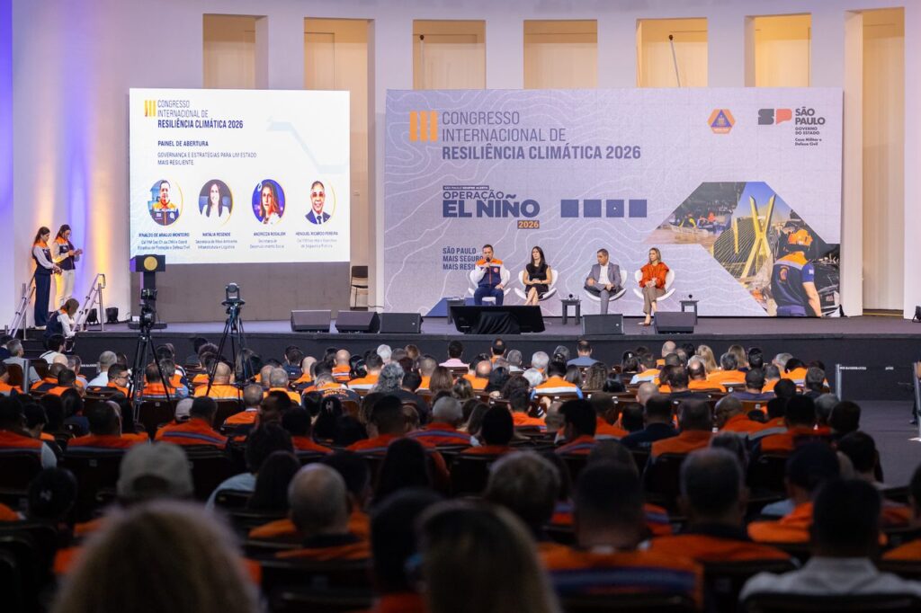 Painel de abertura do III Congresso Internacional de Resiliência Climática 2026 com quatro palestrantes num palco principal perante um auditório lotado, composto maioritariamente por agentes vestindo coletes cor-de-laranja da Defesa Civil.