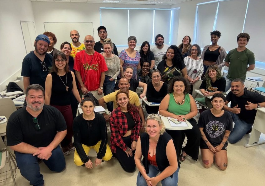 Uma foto de grupo em uma sala de aula. Cerca de 25 pessoas estão reunidas entre carteiras escolares, algumas sentadas e outras em pé, posando para a câmera. O ambiente tem paredes brancas, janelas com persianas fechadas e iluminação interna. A cena sugere um curso, oficina ou atividade de capacitação coletiva.
