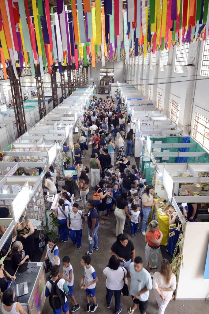 Visão superior do pavilhão repleto de visitantes circulando pelos corredores de stands, com o teto decorado por fitas coloridas penduradas.