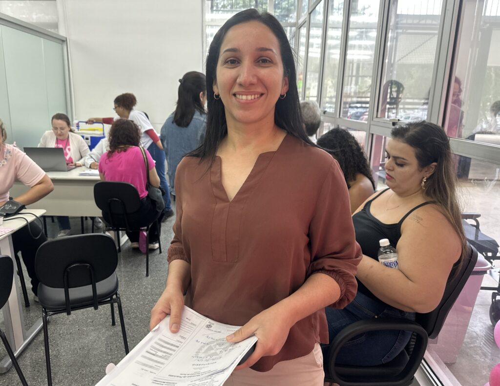 Retrata Eide Lúcio Machado, de 38 anos, servidora da Secretaria de Saúde, segurando documentos de atendimento e destacando a agilidade dos serviços.