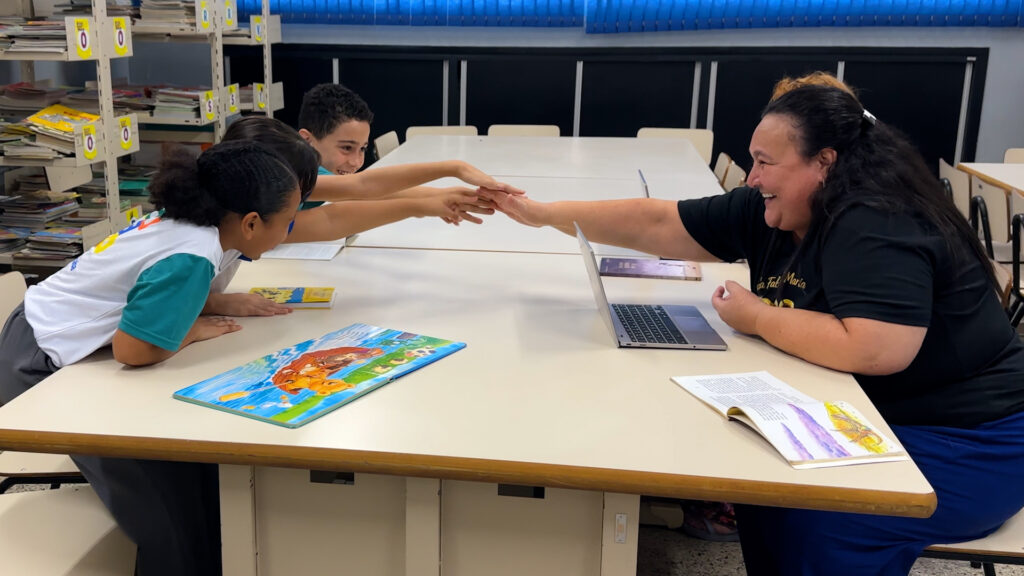 A educadora Sandra Rodrigues de Souza sorri enquanto faz um gesto de cumprimento (high five) com as mãos sobrepostas às de três estudantes numa mesa de biblioteca escolar.