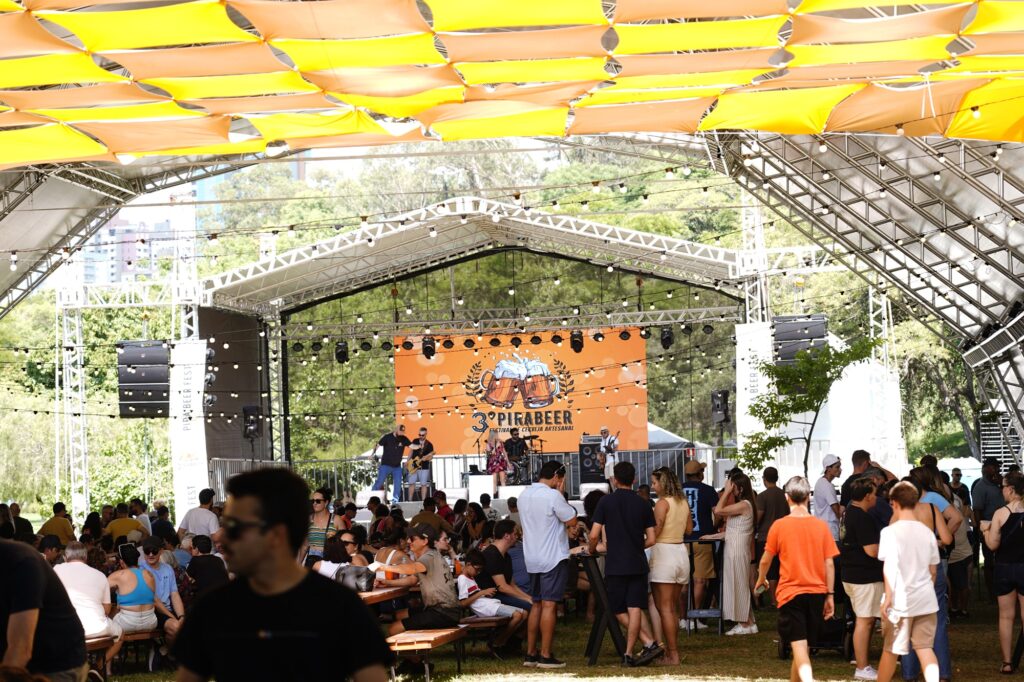 Um festival ao ar livre com música e cerveja está acontecendo sob uma cobertura colorida com muitas pessoas reunidas.

A imagem retrata um vibrante festival ao ar livre, com um palco ao fundo onde uma banda se apresenta para uma multidão. Uma grande tela laranja no palco exibe o logotipo "3º Pirabeer Festival Cerveja Artesanal" com a imagem de duas canecas de cerveja espumante. Vários músicos, incluindo um guitarrista, um baterista e vocalistas, estão no palco. Abaixo do palco, uma multidão de pessoas está sentada em mesas de piquenique e em pé, aproveitando o evento. Há uma atmosfera festiva e animada, com muitas pessoas conversando, rindo e possivelmente consumindo cerveja. A cena é iluminada pela luz natural do dia, com luzes de corda decorativas penduradas sobre a área do público e do palco, adicionando um toque festivo. A cobertura acima é feita de painéis suspensos em tons de amarelo e laranja, criando um ambiente sombreado e colorido. Ao redor do palco e da área do público, há árvores e vegetação, sugerindo que o festival ocorre em um parque ou espaço verde. A multidão é diversificada em termos de idade e vestimenta, com cores variadas em suas roupas. A composição da imagem captura a amplitude do evento, com o palco e a multidão dominando a cena central, e detalhes como as estruturas metálicas do palco e a cobertura colorida adicionando textura e profundidade ao quadro geral. O humor geral é alegre e descontraído, típico de um festival de verão.