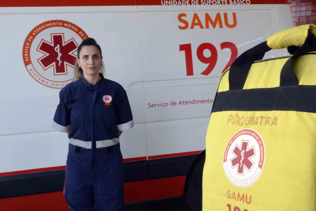 Retrato de plano médio (cortado um pouco acima da cintura) de uma profissional vestida com o uniforme azul-marinho do SAMU. Em primeiro plano, no canto inferior direito, destaca-se parcialmente uma mala de emergência amarela com a identificação "PSIQUIATRA" e o logotipo do serviço, tendo ao fundo uma viatura do SAMU com os números "192".
