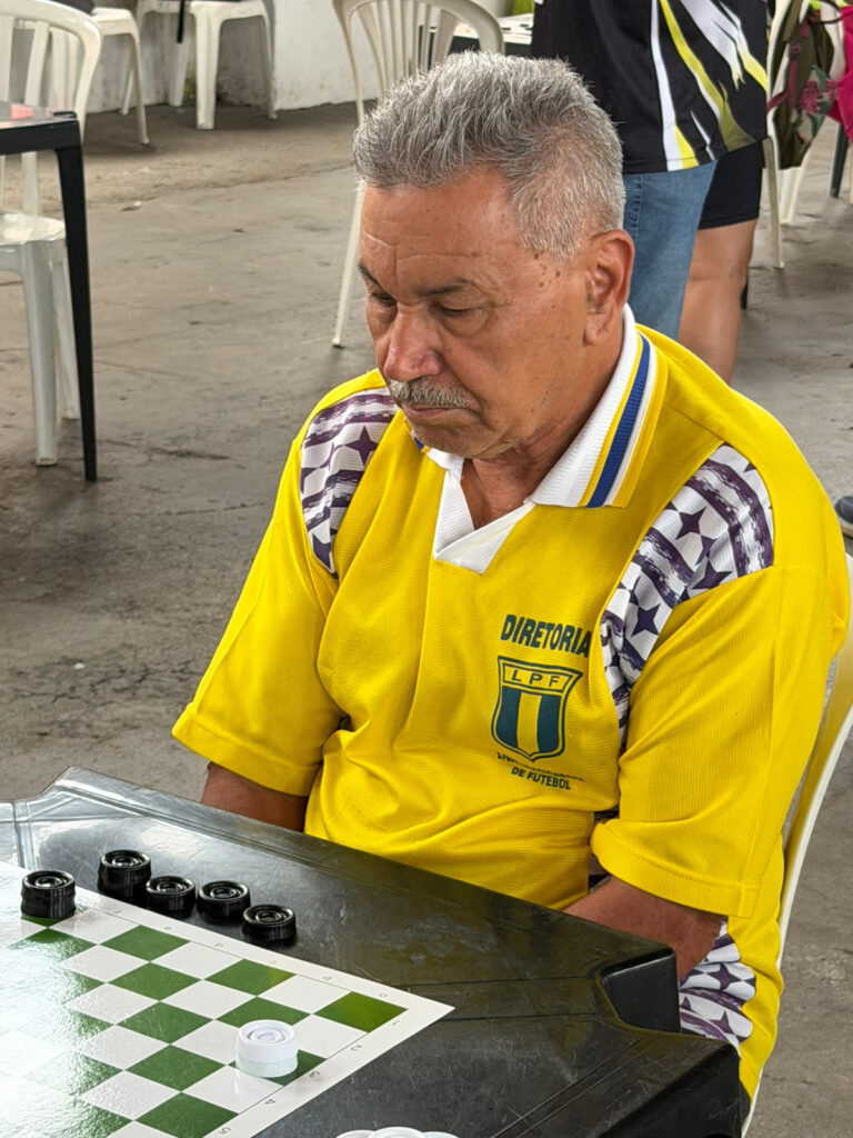 Um homem sénior concentrado a jogar uma partida de damas. Ele está vestido com uma t-shirt amarela da "Diretoria LPF" e encontra-se atento ao tabuleiro com peças pretas e brancas.