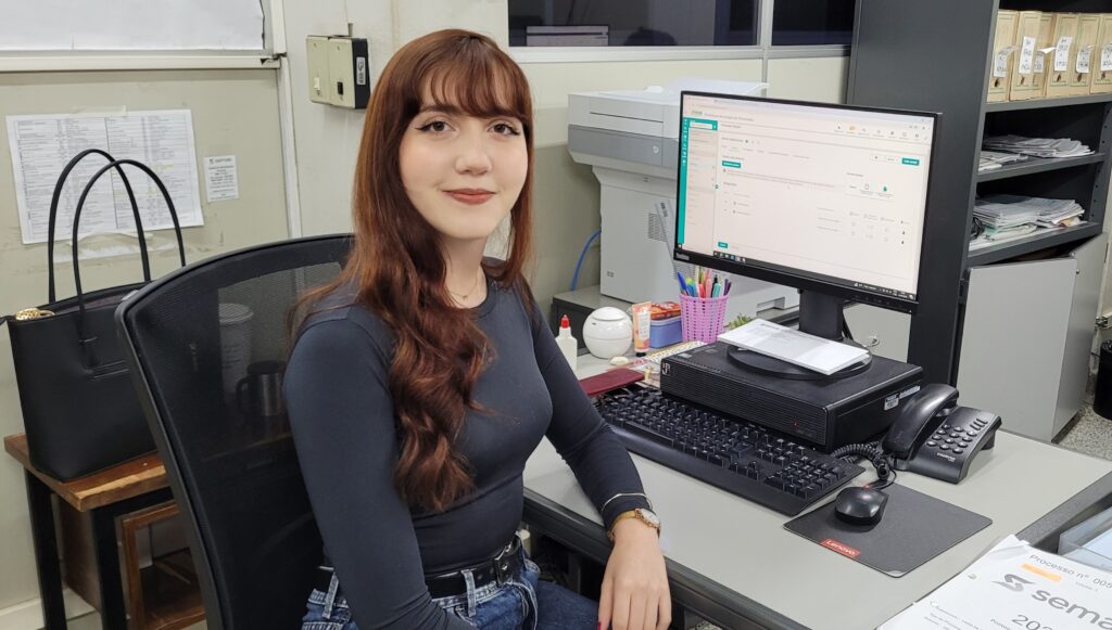 Emily Lambertucci sorri para a câmara sentada à sua secretária, posicionada em frente ao computador e com o sistema do Semae visível no ecrã.