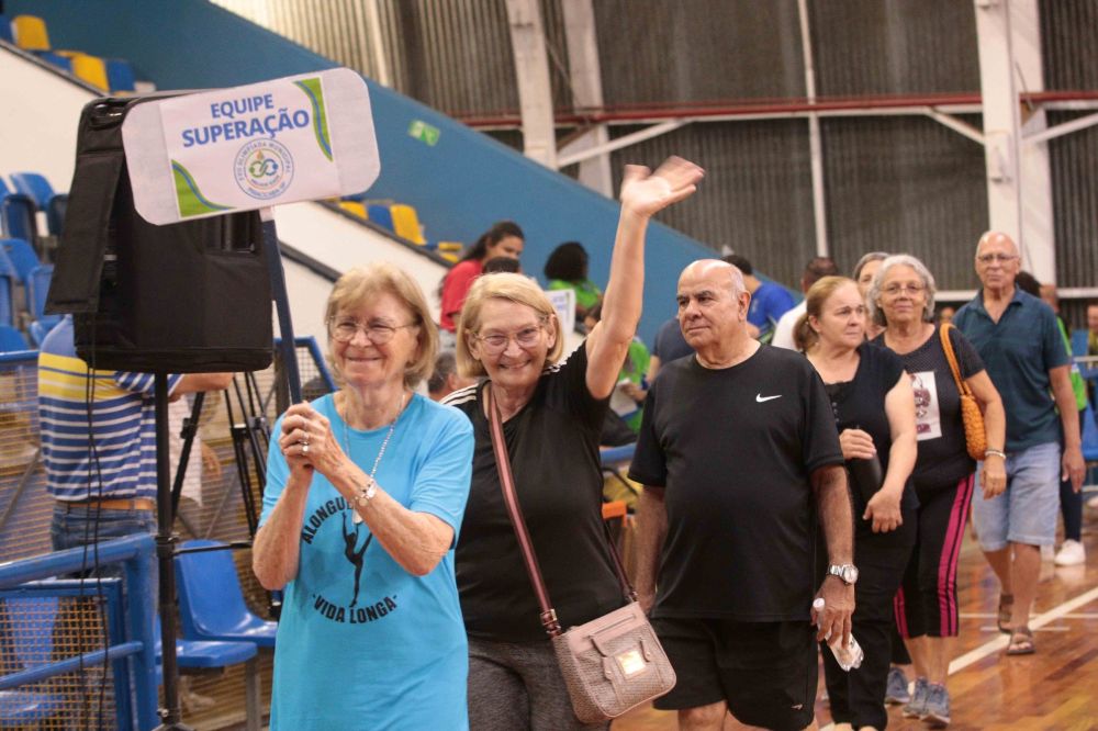 Atletas da melhor idade desfilaram sorridentes na quadra, com uma participante em destaque a acenar alegremente para o público enquanto segura a placa da "Equipe Superação".