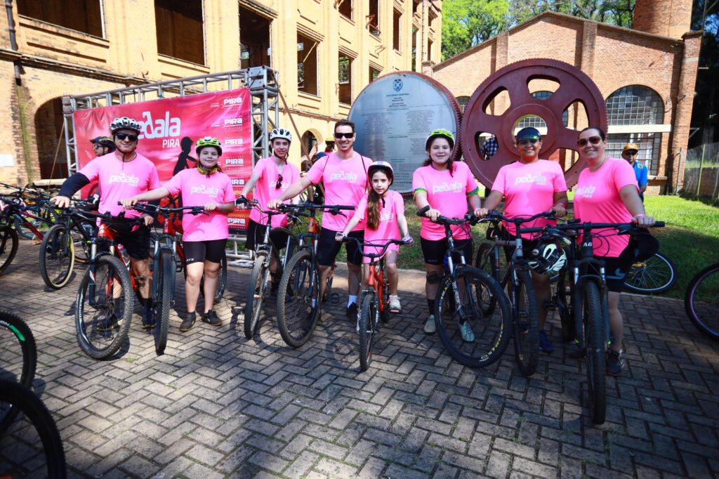 Uma família de ciclistas de camisas cor-de-rosa e capacetes posa com as suas bicicletas em frente a um edifício histórico de tijolos e uma grande engrenagem de ferro no Engenho Central.
