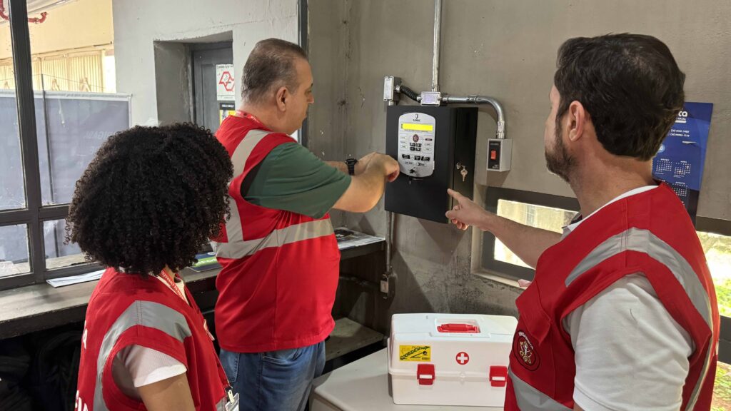 O grupo de brigadistas observa e aponta para o painel central do sistema de alarme de incêndio fixado na parede, ao lado de uma caixa de primeiros socorros.