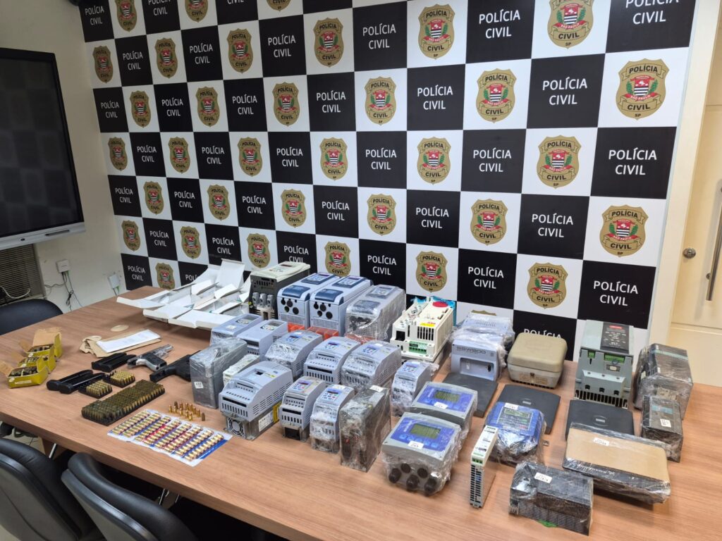 Uma mesa de madeira onde estão dispostos vários equipamentos eletrónicos recuperados (como inversores e controladores industriais), além de armas de fogo, carregadores e grande quantidade de munições, tendo ao fundo uma parede com o logotipo e o padrão quadriculado da Polícia Civil.